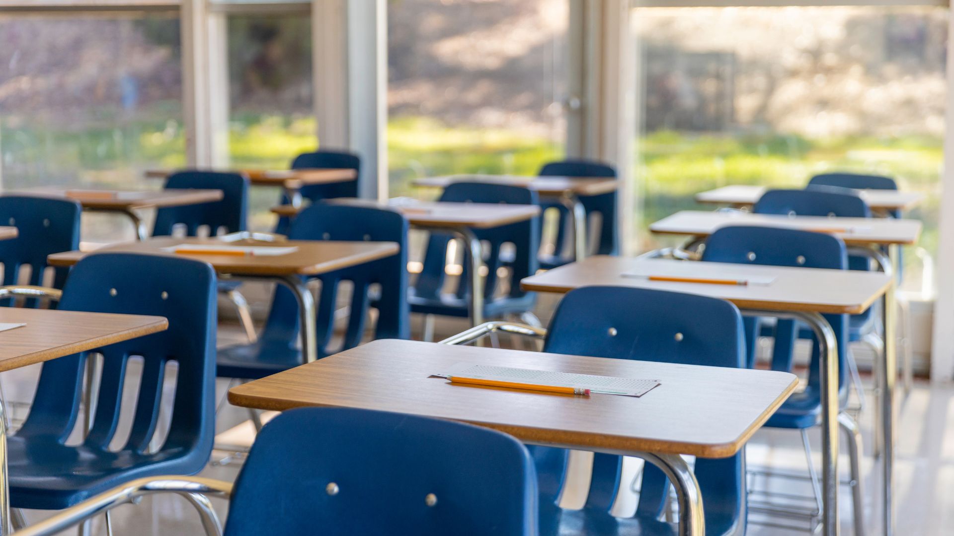 Empty classroom with rows of wooden desks and blue chairs. Pencils and paper sit on each desk. Sunlight streams through large windows, creating a calm atmosphere.