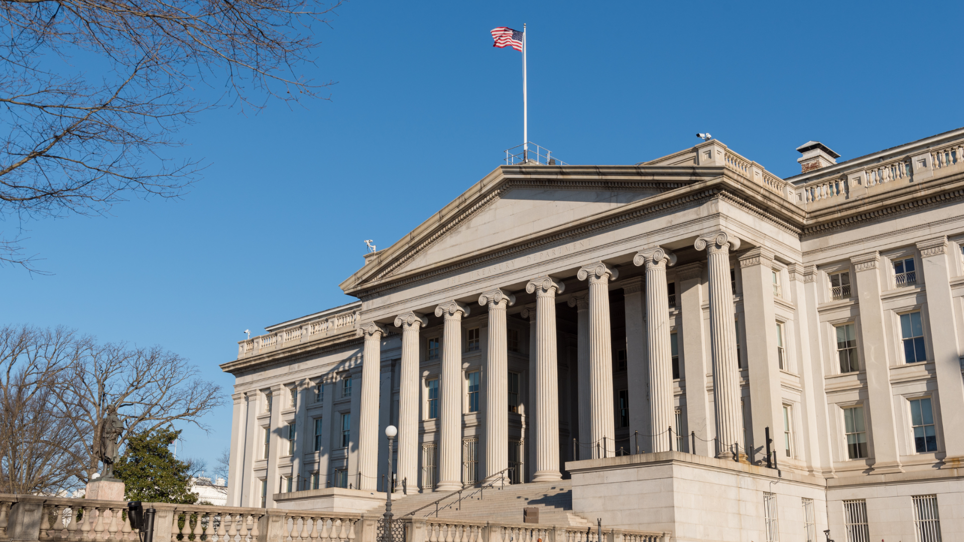 Neoclassical building with tall columns, an American flag flying atop, set against a clear blue sky. Bare trees frame the scene, creating a solemn tone.