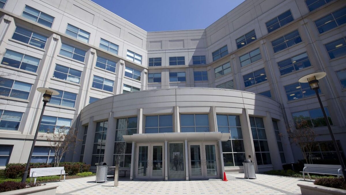 "Modern office building with a curved entrance, large windows, and a light gray facade under a clear blue sky. Benches and lamps flank the entrance."