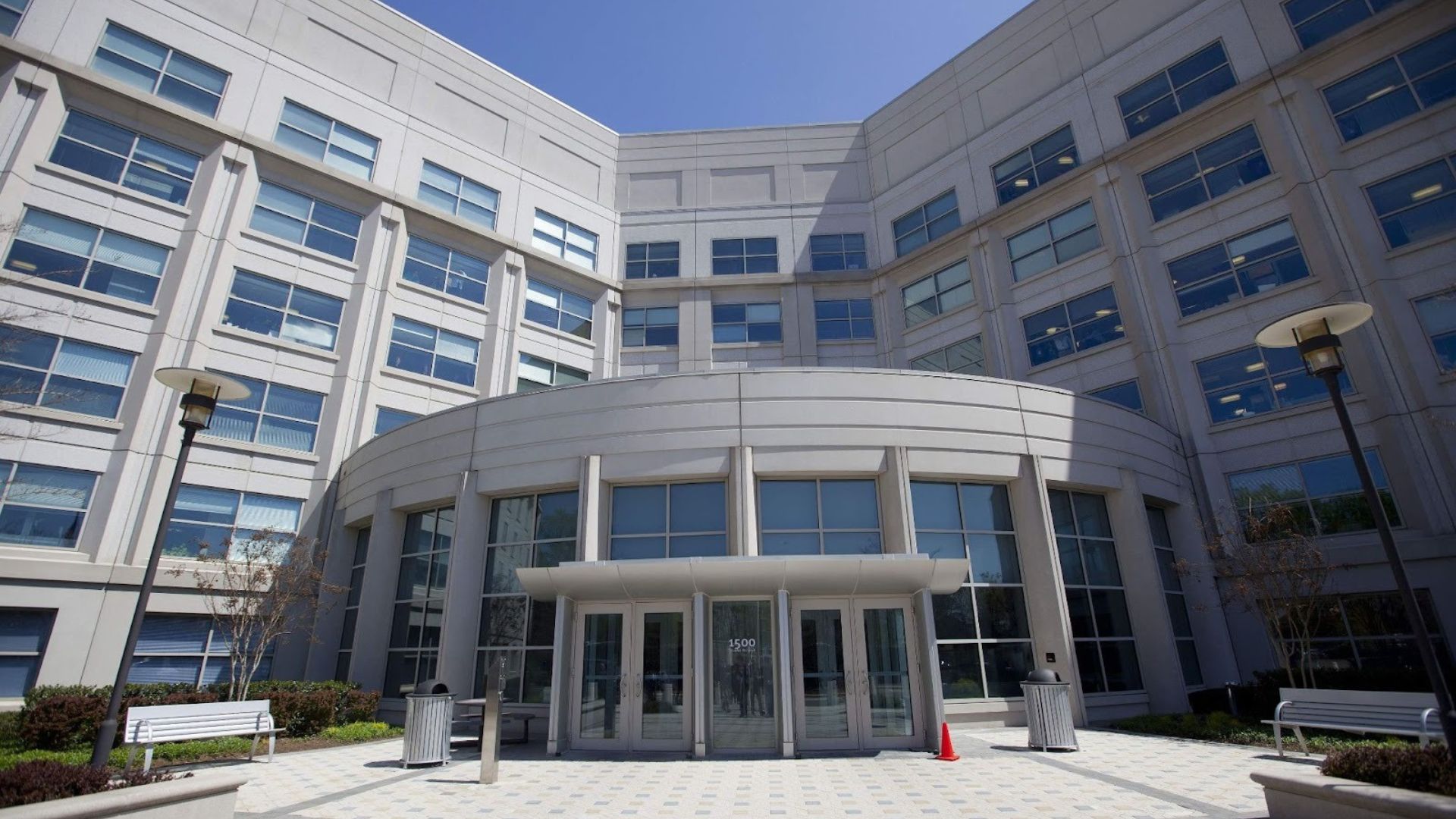 "Modern office building with a curved entrance, large windows, and a light gray facade under a clear blue sky. Benches and lamps flank the entrance."